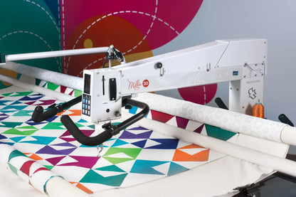 The APQS Demo Millie 30" Long Arm Quilting Machine is quilting a vibrant geometric quilt against an abstract, multicolored background with dotted lines. The white APQS machine features a touchscreen and black handles.