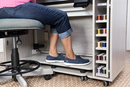A person wearing jeans and navy sneakers sits at a Koala All-In One Embroidery Studio in White and Gray, feet on the footrest; nearby, colorful quilting thread spools are stored on a cabinet shelf.
