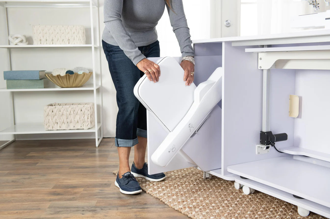 A woman in jeans and sneakers adjusts the white extension panel of a Koala All-In One Embroidery Studio in White and Gray, set in a bright, organized craft room with shelves and quilting storage baskets in the background.