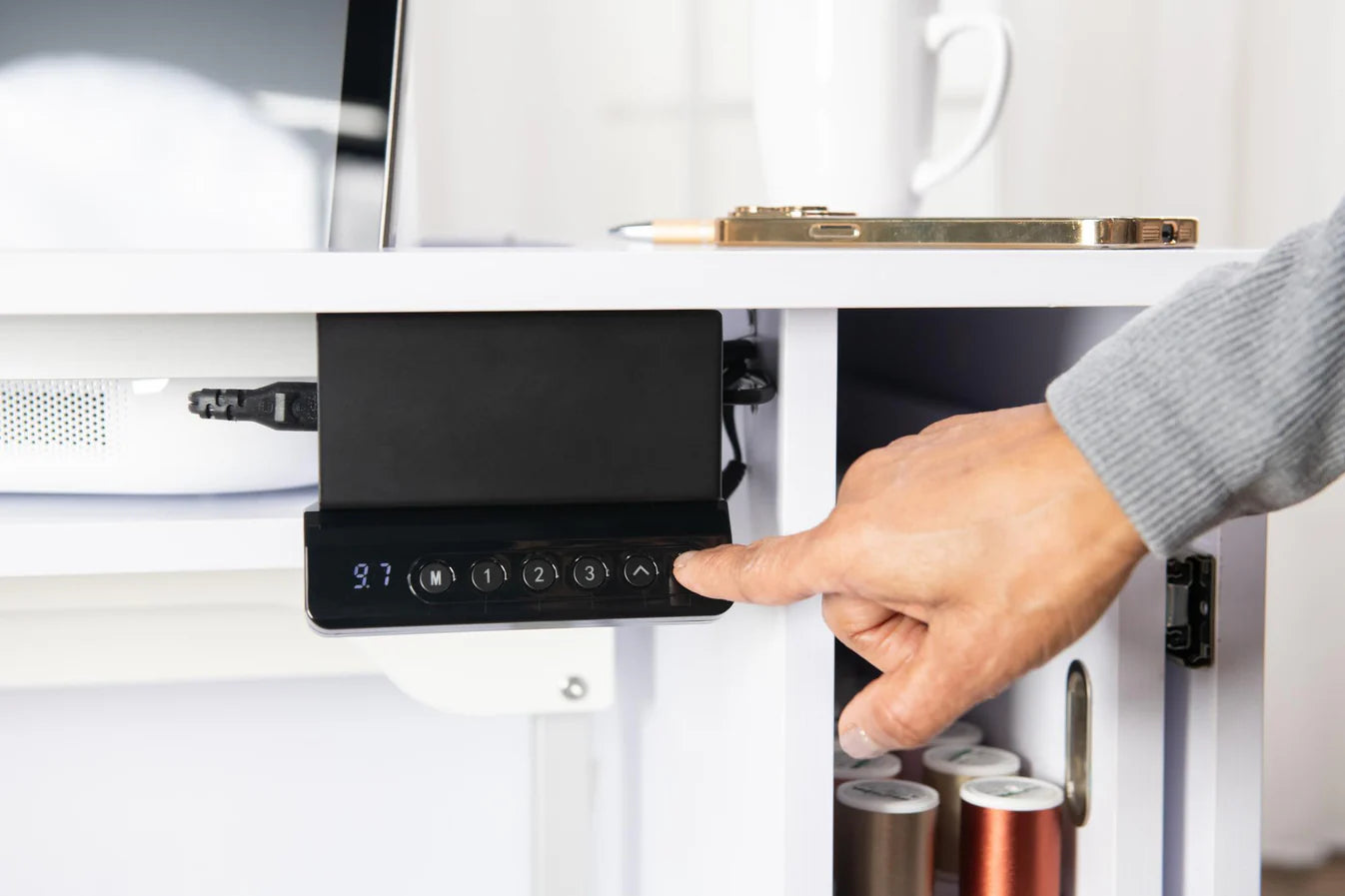 A person presses a button on the Koala All-In One Embroidery Studio in White and Gray, mounted under a sewing cabinet, with a drawer of thread spools and office items like a laptop and notebook nearby.