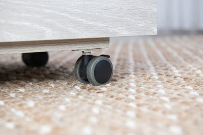 A close-up of a black and gray caster wheel attached to the bottom of a Koala Studios Serger Center in Gray, resting on a beige textured carpet with white accents.