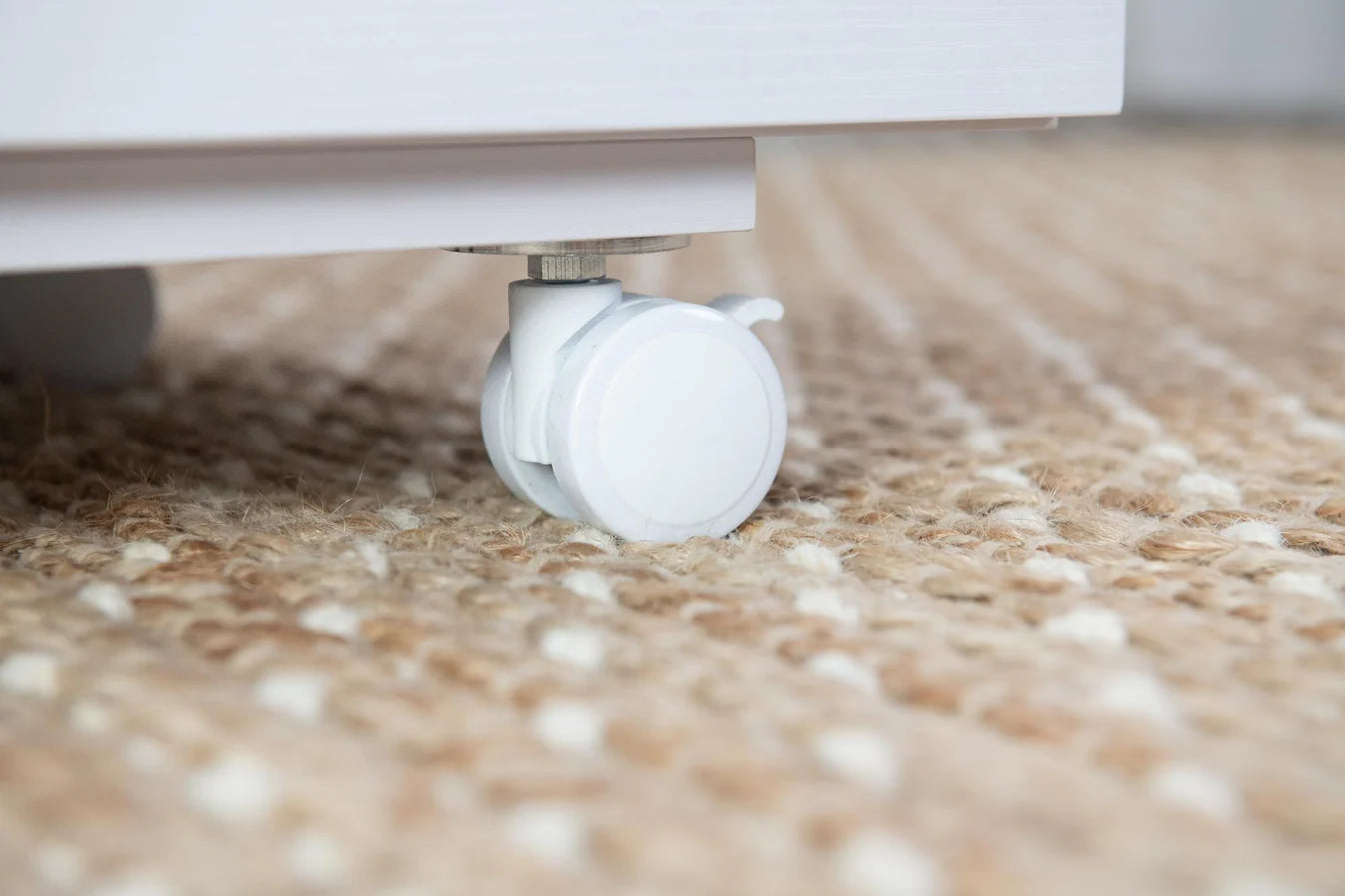Close-up of a white caster wheel attached to the bottom of the Koala Studios Cutting Table in White, resting on a textured beige and cream woven rug.