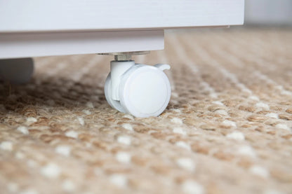 Close-up of a white caster wheel attached to the bottom of the Koala Studios Cutting Table in White, resting on a textured beige and cream woven rug.