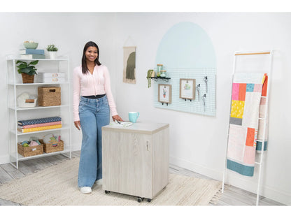 A woman smiles in a bright, organized craft room featuring shelves of supplies, a Koala Compact Sewing Cabinet in White and Gray, a wall pegboard with art, and a colorful quilt displayed on a rack.