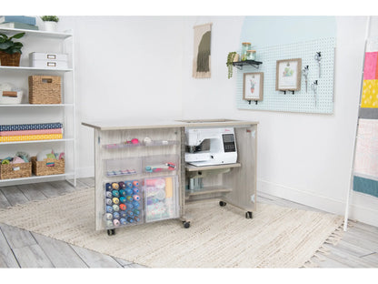 A craft room featuring the Koala Compact Sewing Cabinet in White and Gray, which holds a sewing machine, thread spools, and clear storage boxes. Shelves with baskets, fabric, supplies, plus a pegboard and art enhance the space.