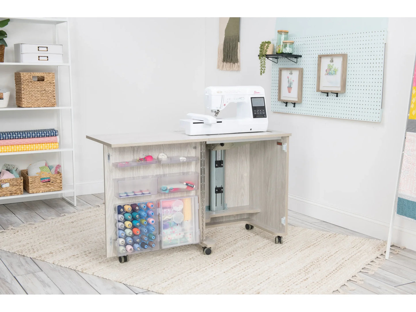 The Koala Compact Sewing Cabinet in White and Gray, featuring a sewing machine on top, storage shelves for thread and supplies below, and wheels for mobility, sits on a beige rug in a bright craft room with wall decor and shelving.