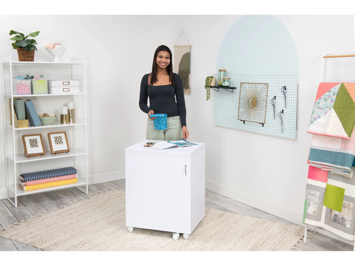 A woman smiles behind the Koala Compact Sewing Cabinet in White and Gray, set on wheels in a bright, organized craft room with shelves, framed art, hanging fabrics, and crafting supplies.