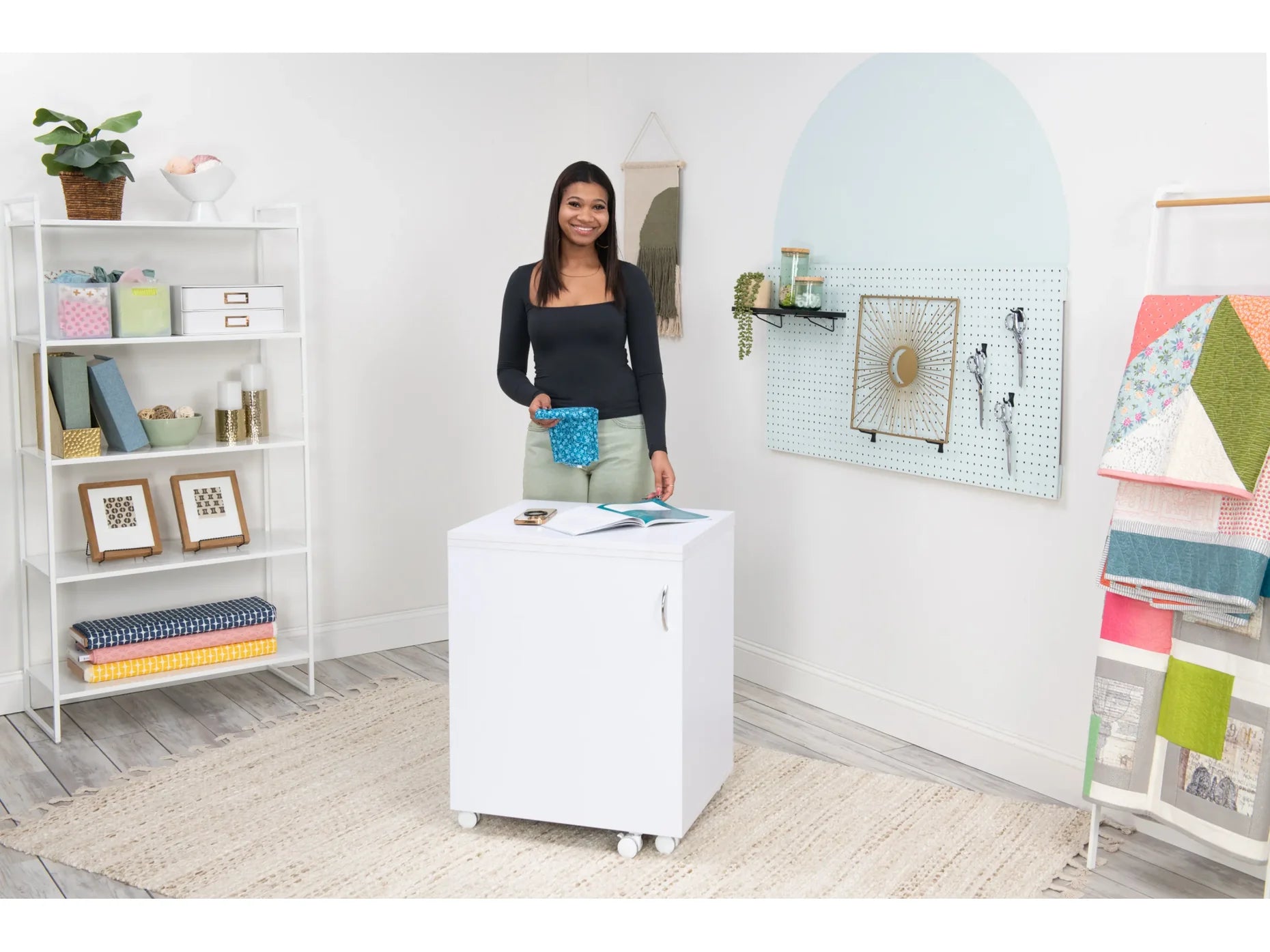 A woman smiles behind the Koala Compact Sewing Cabinet in White and Gray, set on wheels in a bright, organized craft room with shelves, framed art, hanging fabrics, and crafting supplies.