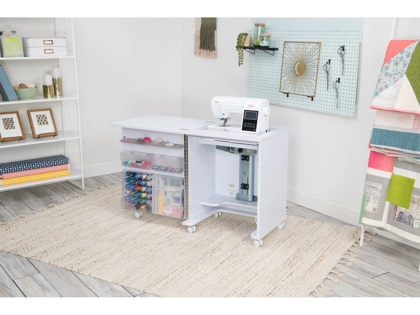 A Koala Compact Sewing Cabinet in White and Gray holds a sewing machine and transparent storage drawers filled with supplies in a bright craft room with shelves, a pegboard, and colorful quilts on display.