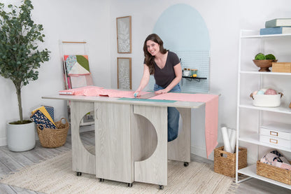 A woman trims pink fabric with a rotary cutter at the Koala Studios Cutting Table in white, surrounded by an organized crafting center with shelves, baskets, supplies, a potted tree, and a cozy rug.