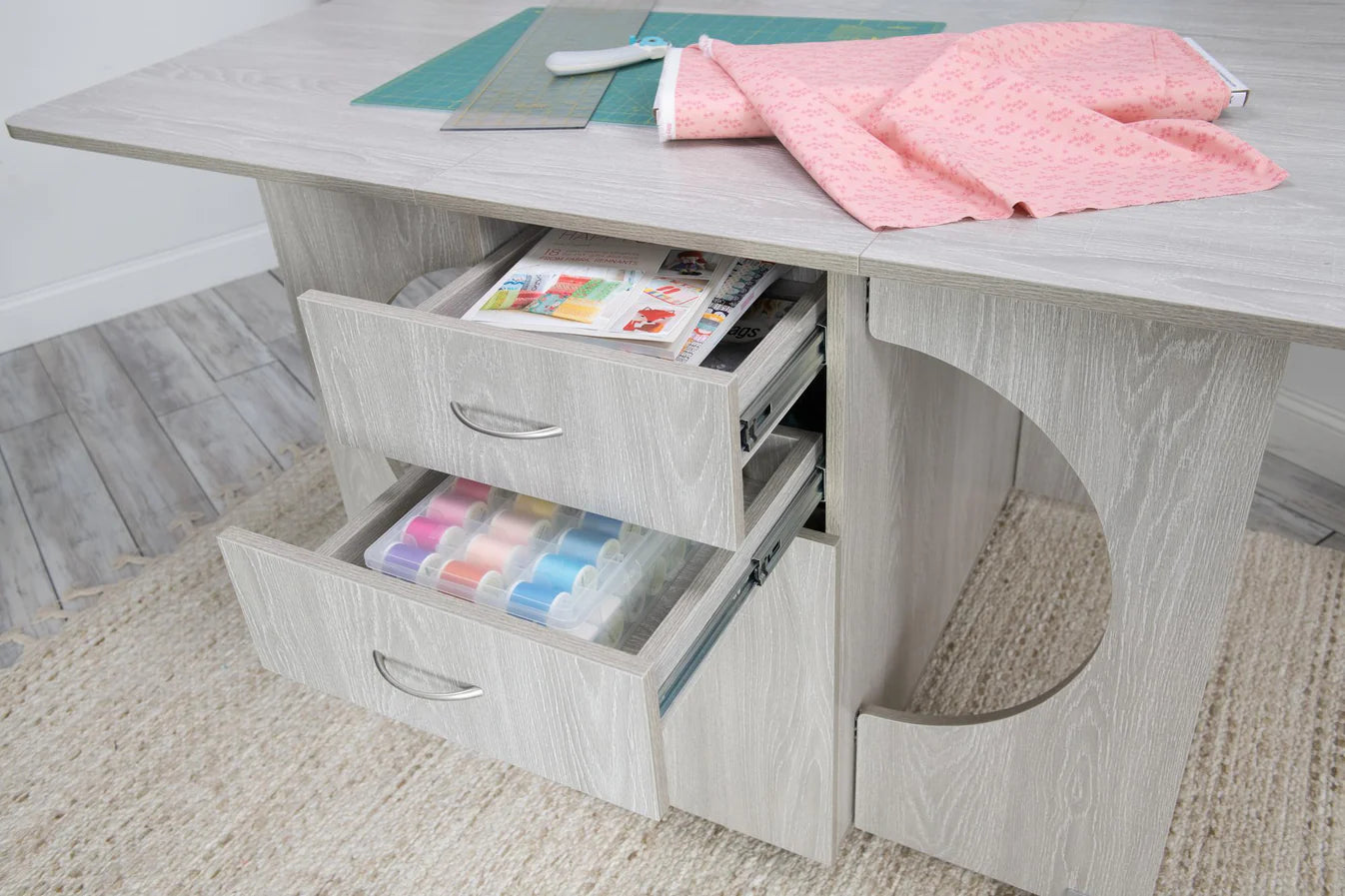 The Koala Studios Cutting Table in White or Gray features two open drawers for threads and magazines, with space on top for a rotary cutter, ruler, and fabric. It sits atop a textured beige rug—ideal for any crafting center.