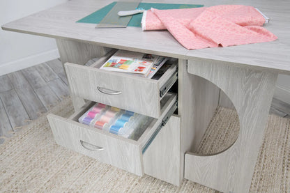 The Koala Studios Cutting Table in White or Gray features two open drawers for threads and magazines, with space on top for a rotary cutter, ruler, and fabric. It sits atop a textured beige rug—ideal for any crafting center.