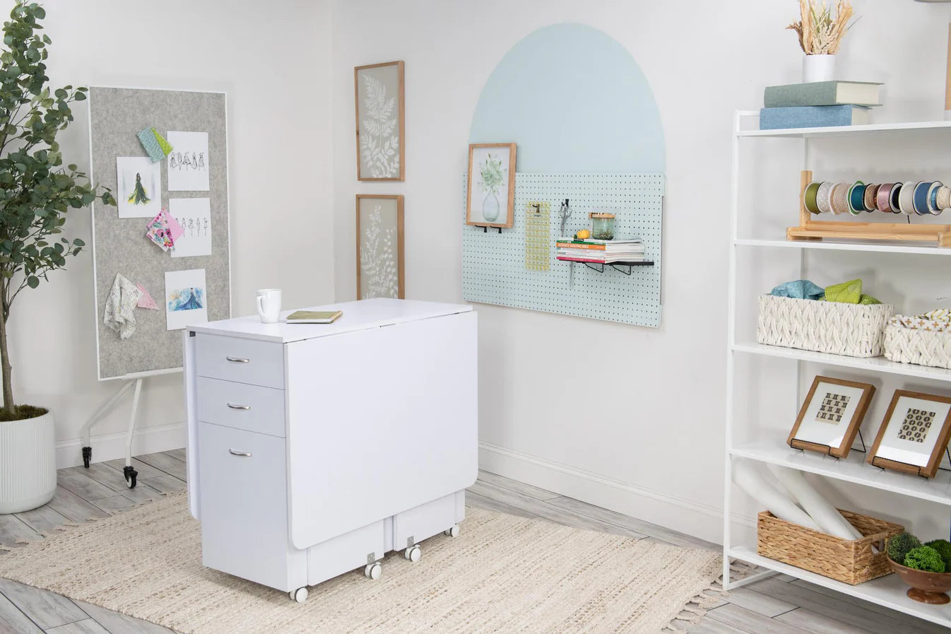 A bright, organized crafting center features the Koala Studios Cutting Table in White or Gray, pegboard on the wall, ribbon and art supply shelves, a corkboard with sketches, a potted plant, and framed art on light walls.