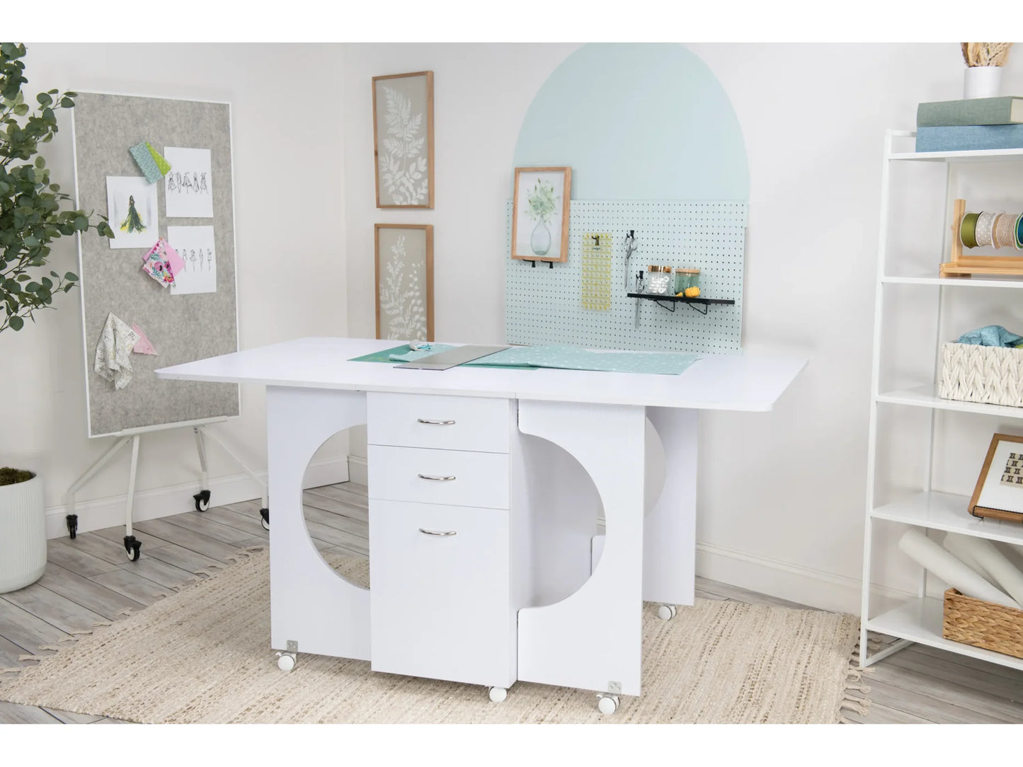 A minimal, organized crafting center with a Koala Studios Cutting Table in White, featuring circular cutouts and wheeled drawers, plus a pegboard with art supplies, cork board for notes, and shelves with baskets.