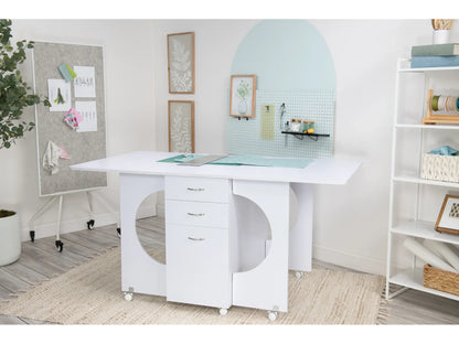 A minimal, organized crafting center with a Koala Studios Cutting Table in White, featuring circular cutouts and wheeled drawers, plus a pegboard with art supplies, cork board for notes, and shelves with baskets.