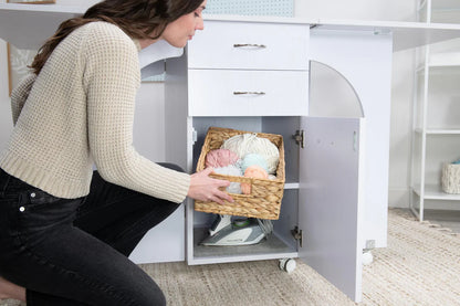 A woman kneels by her Koala Studios Cutting Table in white, placing a basket of yarn onto a lower shelf with the cabinet door open, revealing an iron inside.