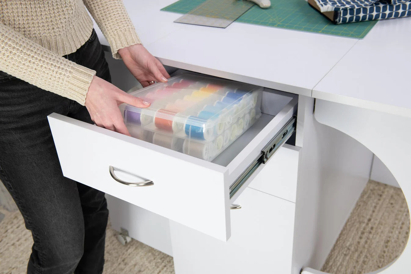 A person places a plastic organizer with colorful thread spools into a Koala Studios Cutting Table in white at their sewing workstation.