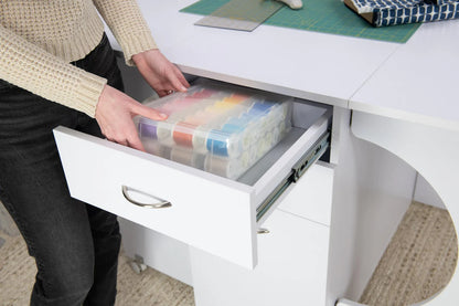 A person places a plastic organizer with colorful thread spools into a Koala Studios Cutting Table in white at their sewing workstation.