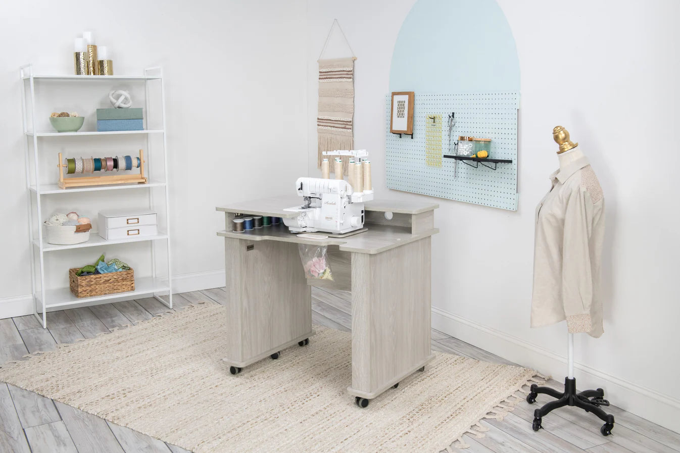 A bright sewing room features the Koala Studios Serger Center in white, holding a serger, shelves with storage for ribbons and supplies, a dress form with a beige shirt, a pegboard for tools, and a woven rug on the light wood floor.