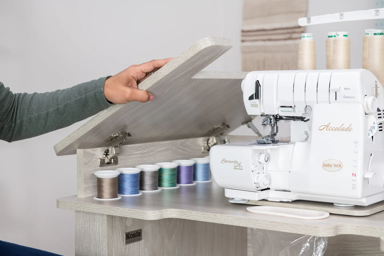 A person lifts the lid of a Koala Studios Serger Center in white, revealing colorful thread spools and a Baby Lock Accolade serger—ideal for organized sewing storage.