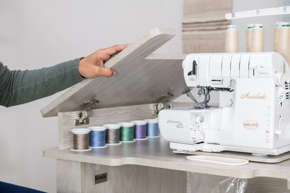 A person lifts the lid of a Koala Studios Serger Center in white, revealing colorful thread spools and a Baby Lock Accolade serger—ideal for organized sewing storage.