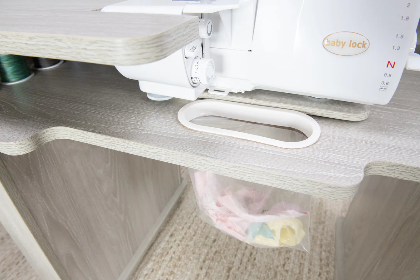 A Baby Lock sewing machine sits on the Koala Studios Serger Center in white, featuring a built-in oval cutout for scraps that leads to a clear plastic bag below—perfect for efficient waste collection and organized sewing.