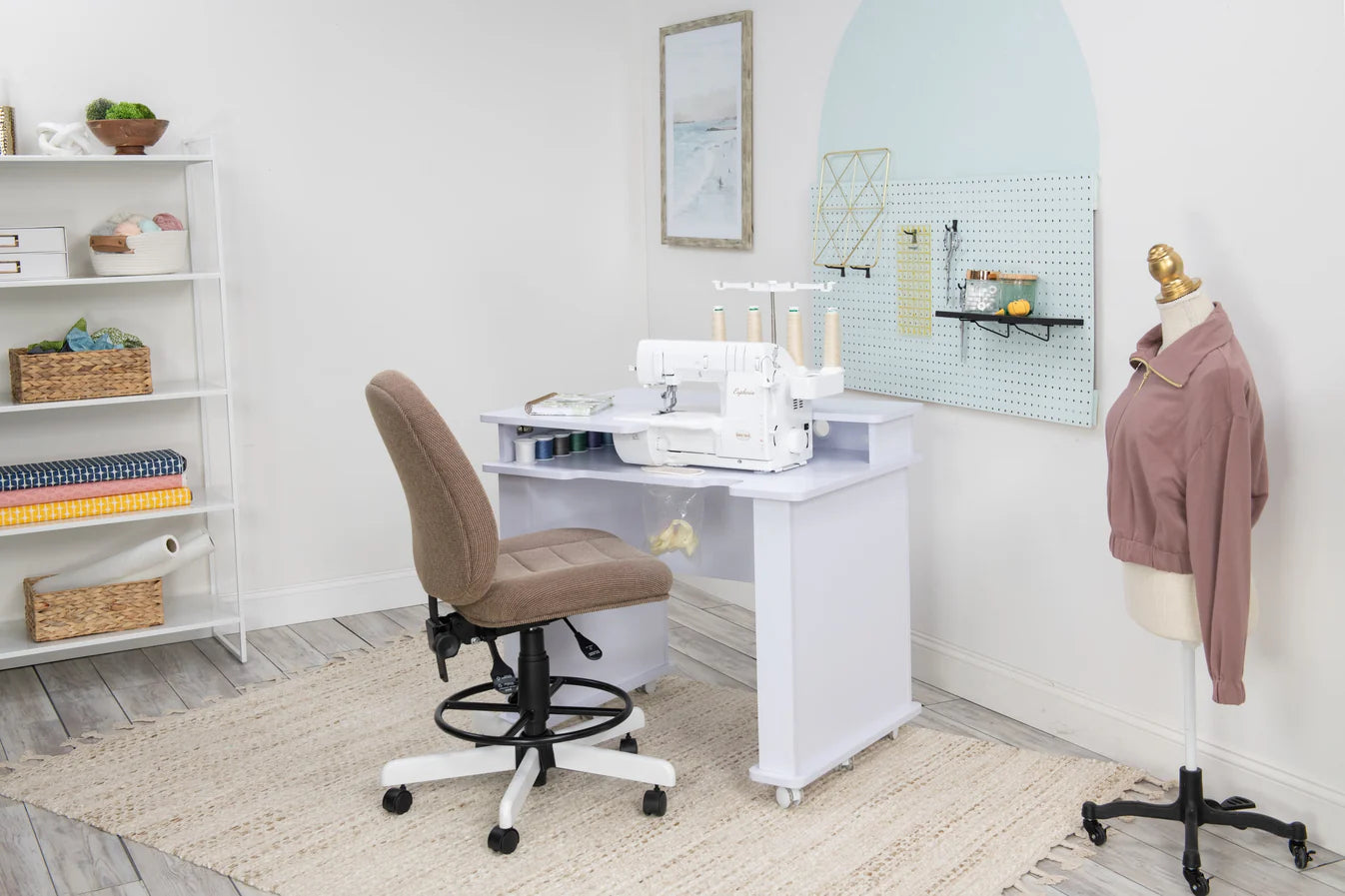 A sewing room features the Koala Studios Serger Center in white holding a sewing machine, a tan office chair, a mannequin with a pink jacket, wall pegboard for tools, and shelves storing fabric, yarn, and baskets.