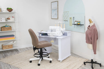 A sewing room features the Koala Studios Serger Center in white holding a sewing machine, a tan office chair, a mannequin with a pink jacket, wall pegboard for tools, and shelves storing fabric, yarn, and baskets.