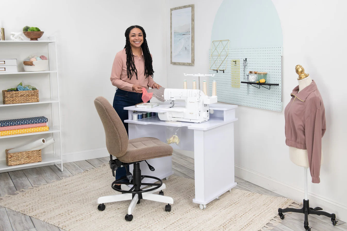 A woman smiles at her Koala Studios Serger Center in white or gray, holding fabric at a sewing desk. The room features shelves for storage, a mannequin with a pink jacket, and neatly arranged sewing supplies on the wall.