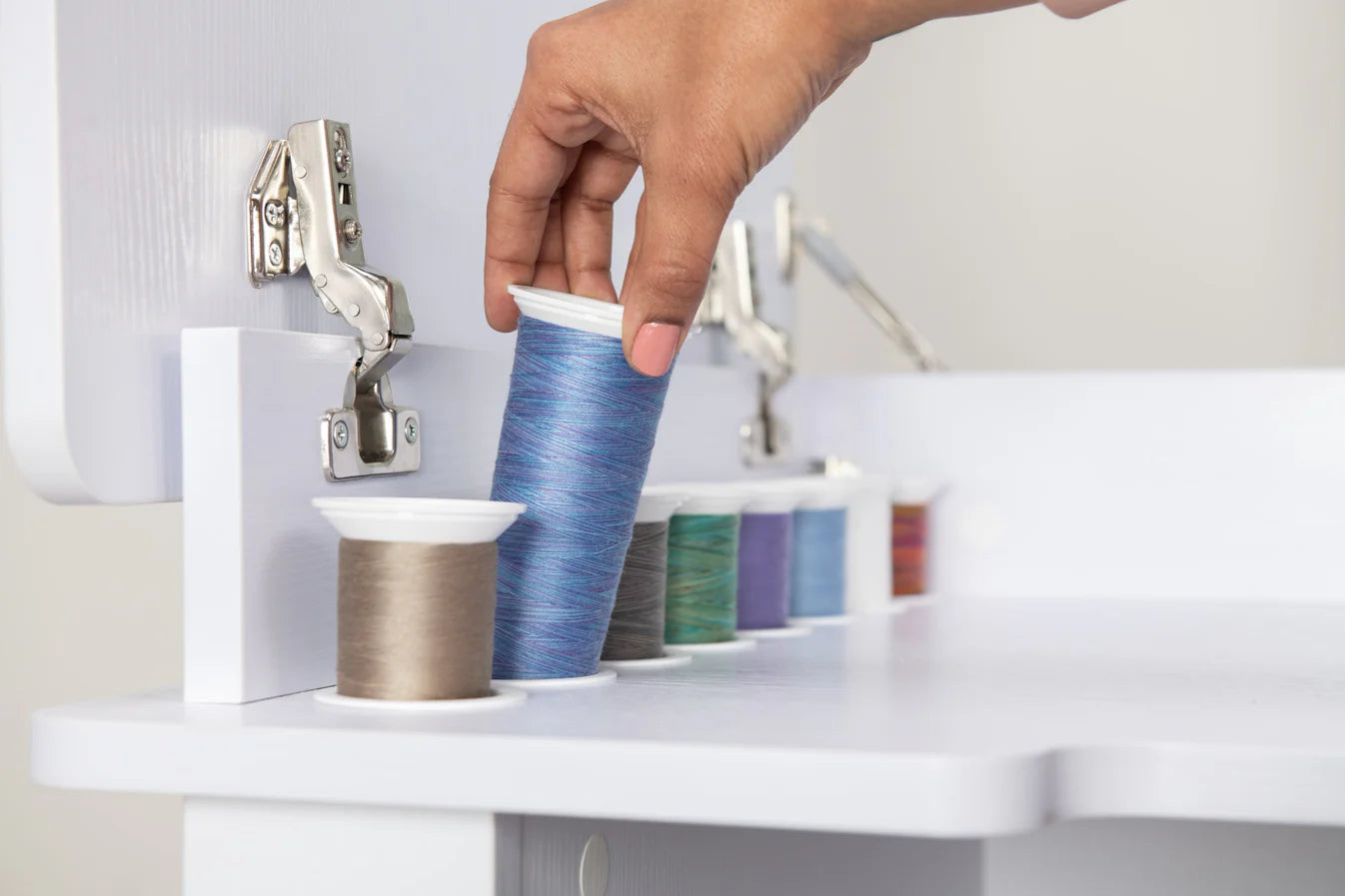A hand selects a spool of blue thread from a row of colorful spools on the Koala Studios Serger Center, available in White or Gray—ideal sewing storage for any craft room.