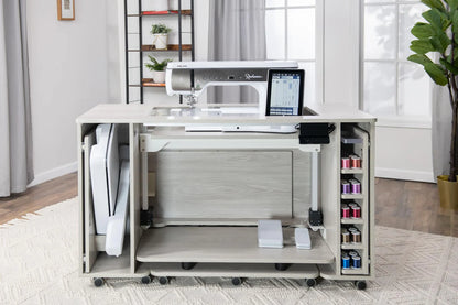 The Koala All-In One Embroidery Studio in White and Gray offers a sleek sewing machine table, open shelves for colorful threads, a folding workspace, and a modern design, perfect for bright rooms with large windows and indoor plants.