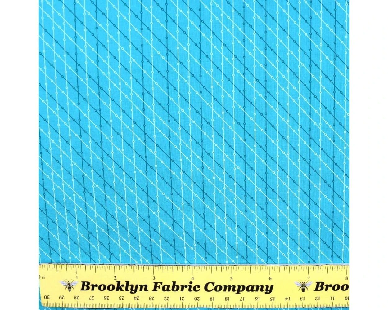 April Showers Water Fabric Yardage by Benartex, featuring diagonal dashed lines in light blue and yellow, is shown with a yellow Brooklyn Fabric Company ruler (about 11 inches) for scale—perfect for fabric projects.