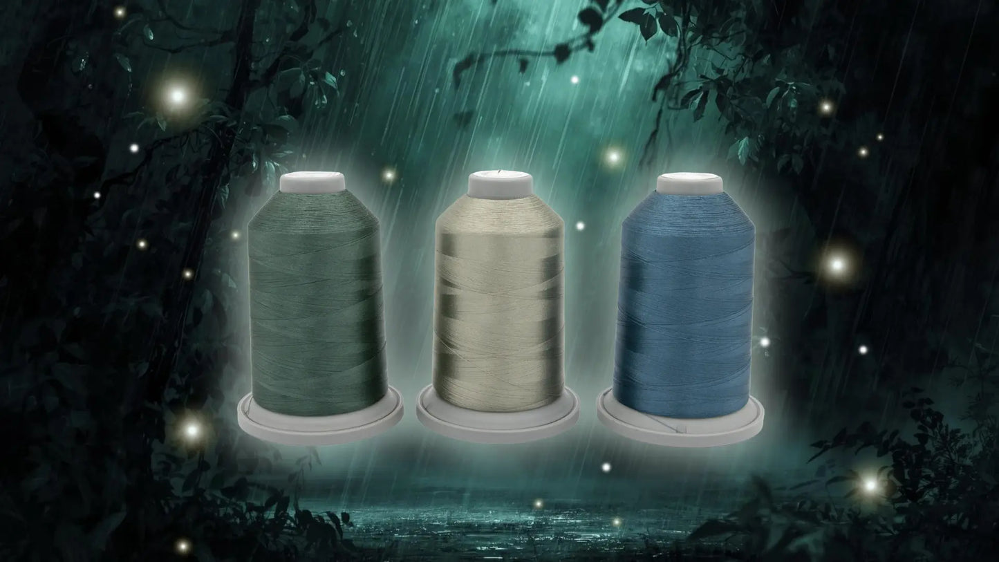 The Stormy Sprite Glide Thread of the Month Bundle by Hab+Dash by Fil Tec features three spools of quilting thread in green, beige, and blue set against a mystical forest backdrop with rain, glowing fireflies, and tree silhouettes.