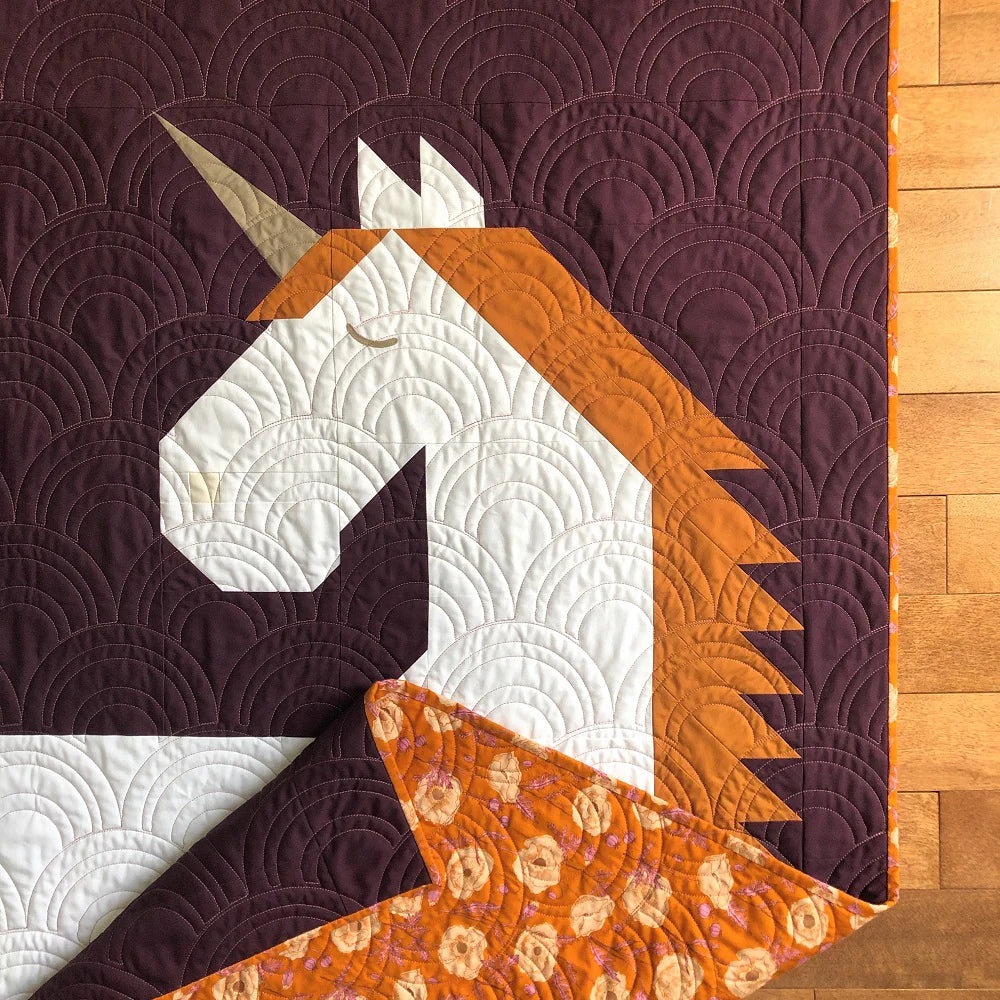 The Unicorn Garden Quilt Pattern by Apples & Beavers features a whimsical white, gold, and brown unicorn head on purple with curved stitching. Corner folds reveal an orange backing with a circular floral print.