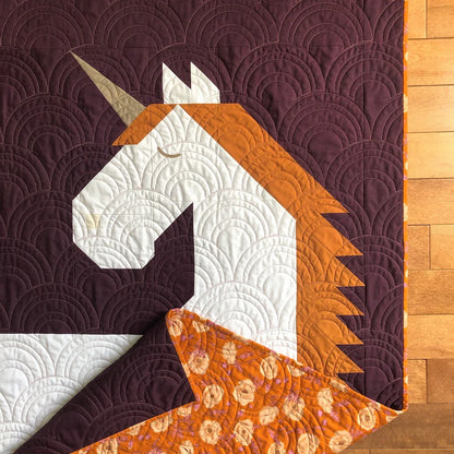 The Unicorn Garden Quilt Pattern by Apples & Beavers features a whimsical white, gold, and brown unicorn head on purple with curved stitching. Corner folds reveal an orange backing with a circular floral print.