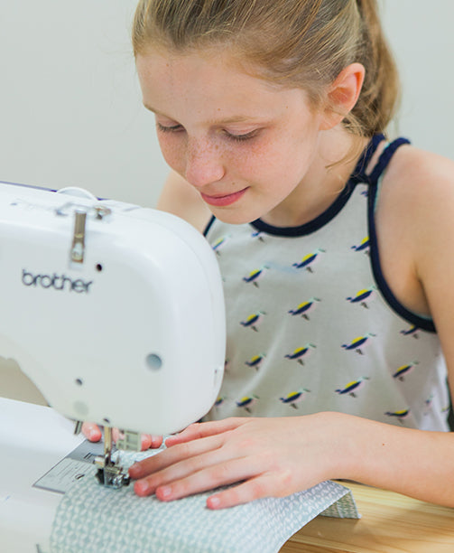 A young girl in a bird-patterned sleeveless top uses a Brother sewing machine to stitch light blue patterned fabric, concentrating on her work.