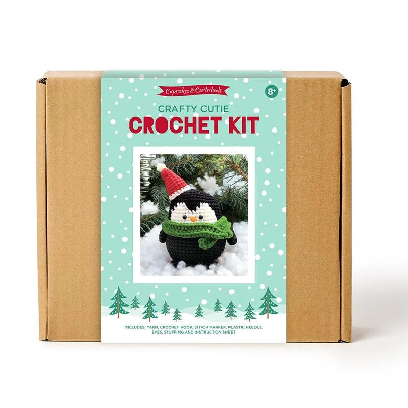 A cardboard box features a United Notions Crafty Cutie Penguin Crochet Kit label, showing a crocheted penguin in a Santa hat amid snow. Suitable for ages 8+, the kit lets you make an adorable handmade plushie.