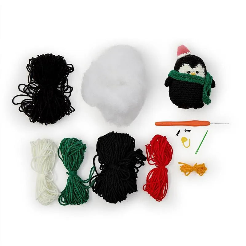 The Crafty Cutie Penguin Crochet Kit by United Notions includes yarn, stuffing, a crochet hook, needles, and all supplies needed to make a handmade penguin plushie wearing a green scarf and Santa hat—perfect for the holidays.