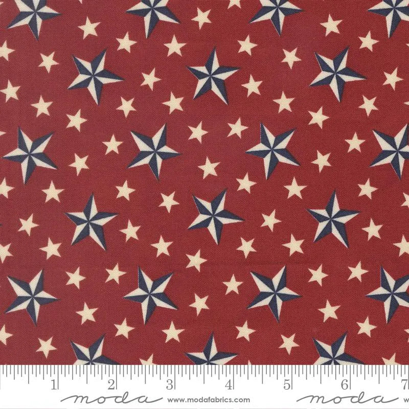 America 250 Years Crimson Tossed Stars Fabric Yardage by United Notions features red fabric with scattered large blue and white stars and small white stars, ideal for patriotic quilts or America 250 celebrations. Ruler at bottom shows inches.