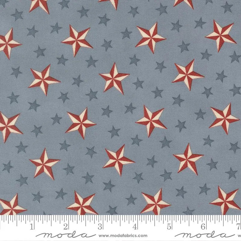 America 250 Years Steel Blue Tossed Stars Fabric Yardage by United Notions features large red, white, and small dark blue stars on a gray background. Ideal for quilts celebrating America's 250th anniversary. Includes inch measurements along the bottom edge.