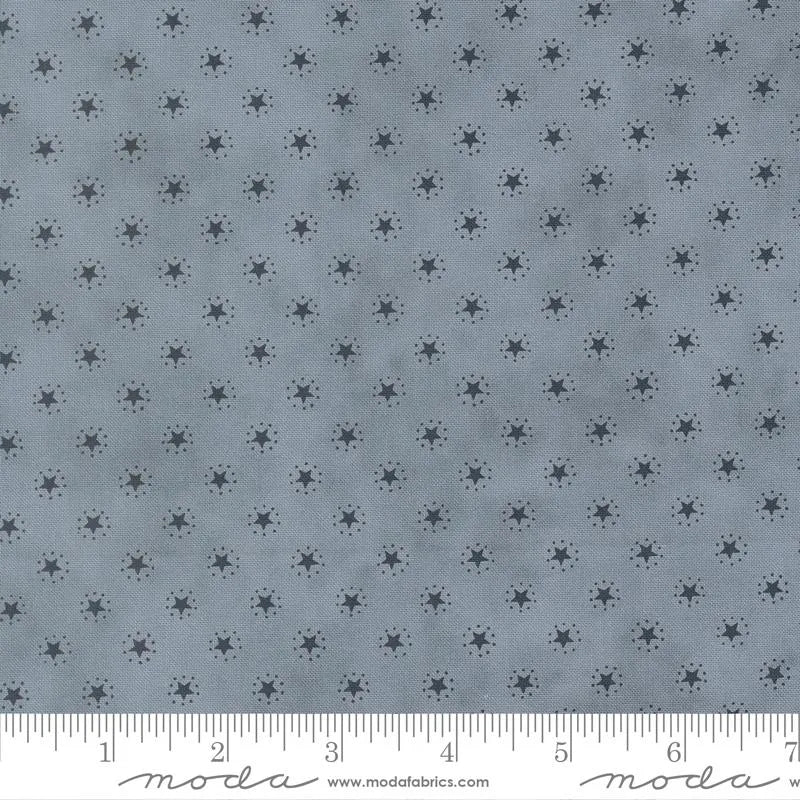 America 250 Years Steel Blue Starbright Blenders Fabric Yardage by United Notions features a gray fabric with small, dark starburst shapes. The inch ruler shows scale, making it ideal as a steel blue blender for patriotic quilt projects.