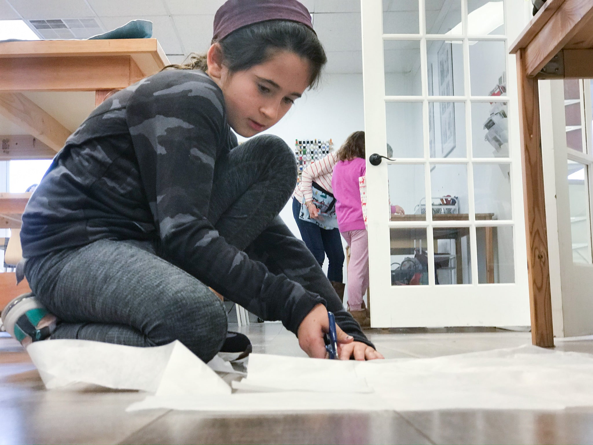 A young girl kneels on the floor, focused on cutting white paper with scissors. She wears a dark outfit and headband. In the background, two other children stand near a glass door in a bright room.