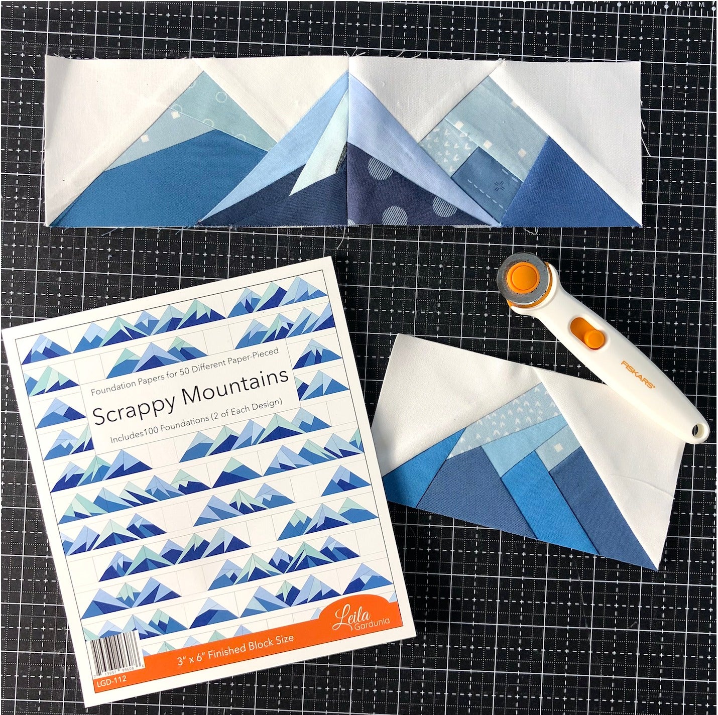 Three blue and white mountain quilt blocks made with the Scrappy Mountains Foundation Paper Piecing Pad by Checker Distributor are displayed on a black grid mat, alongside a rotary cutter and the pad’s instruction booklet.
