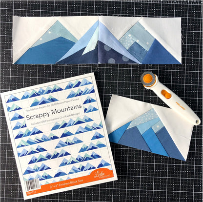 Three blue and white mountain quilt blocks made with the Scrappy Mountains Foundation Paper Piecing Pad by Checker Distributor are displayed on a black grid mat, alongside a rotary cutter and the pad’s instruction booklet.