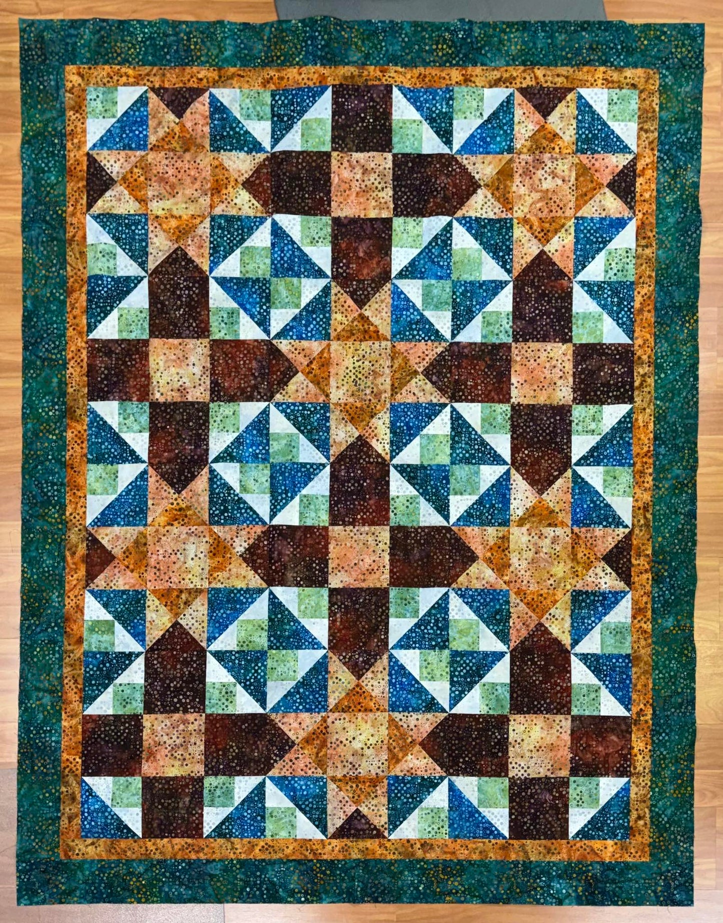 The Luau Torches Mystery Five-0 Quilt by quiltedjoy features triangles and squares in green, blue, brown, and orange fabrics with a dark green border, beautifully displayed on a wooden floor.