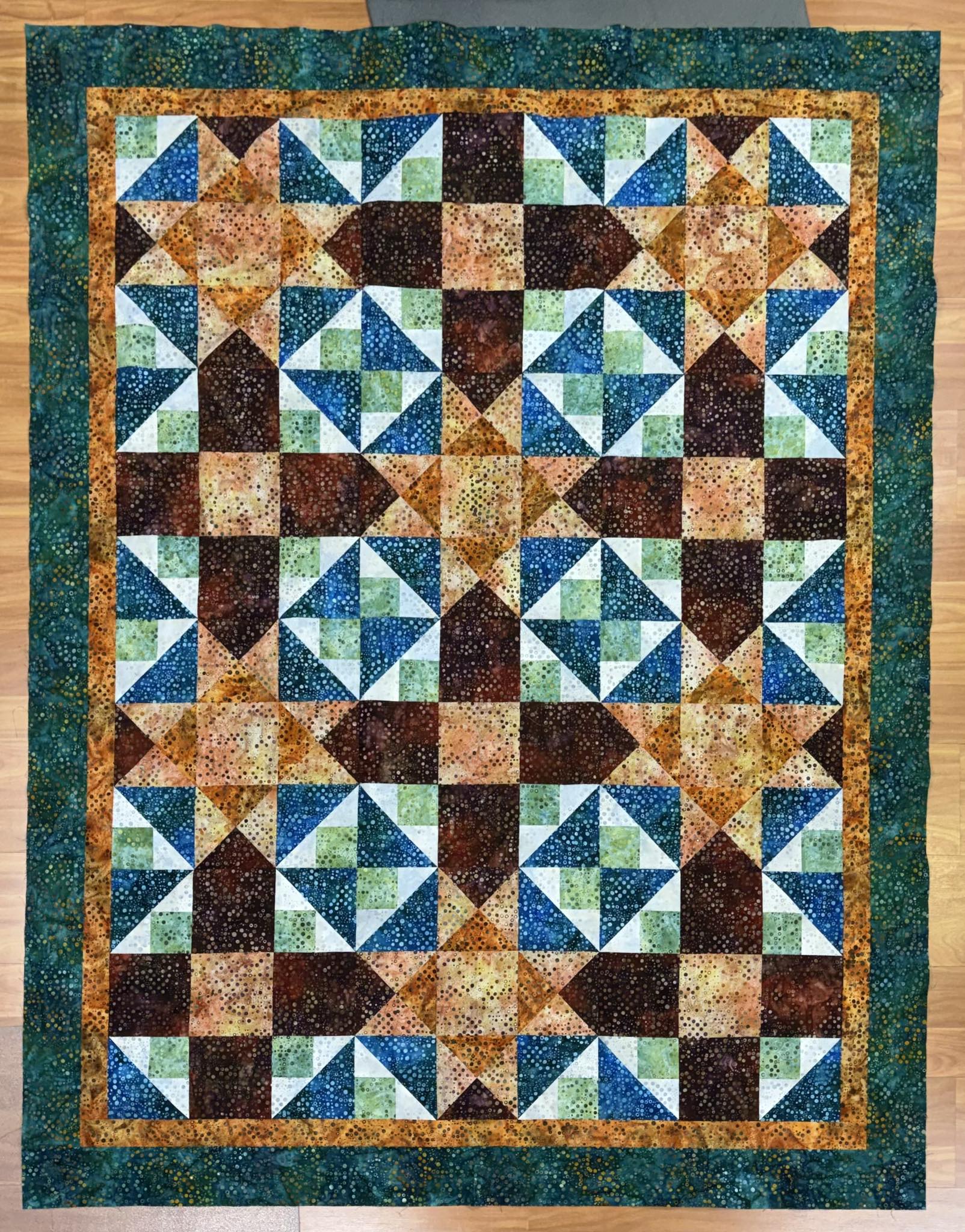 The Luau Torches Mystery Five-0 Quilt by quiltedjoy features triangles and squares in green, blue, brown, and orange fabrics with a dark green border, beautifully displayed on a wooden floor.