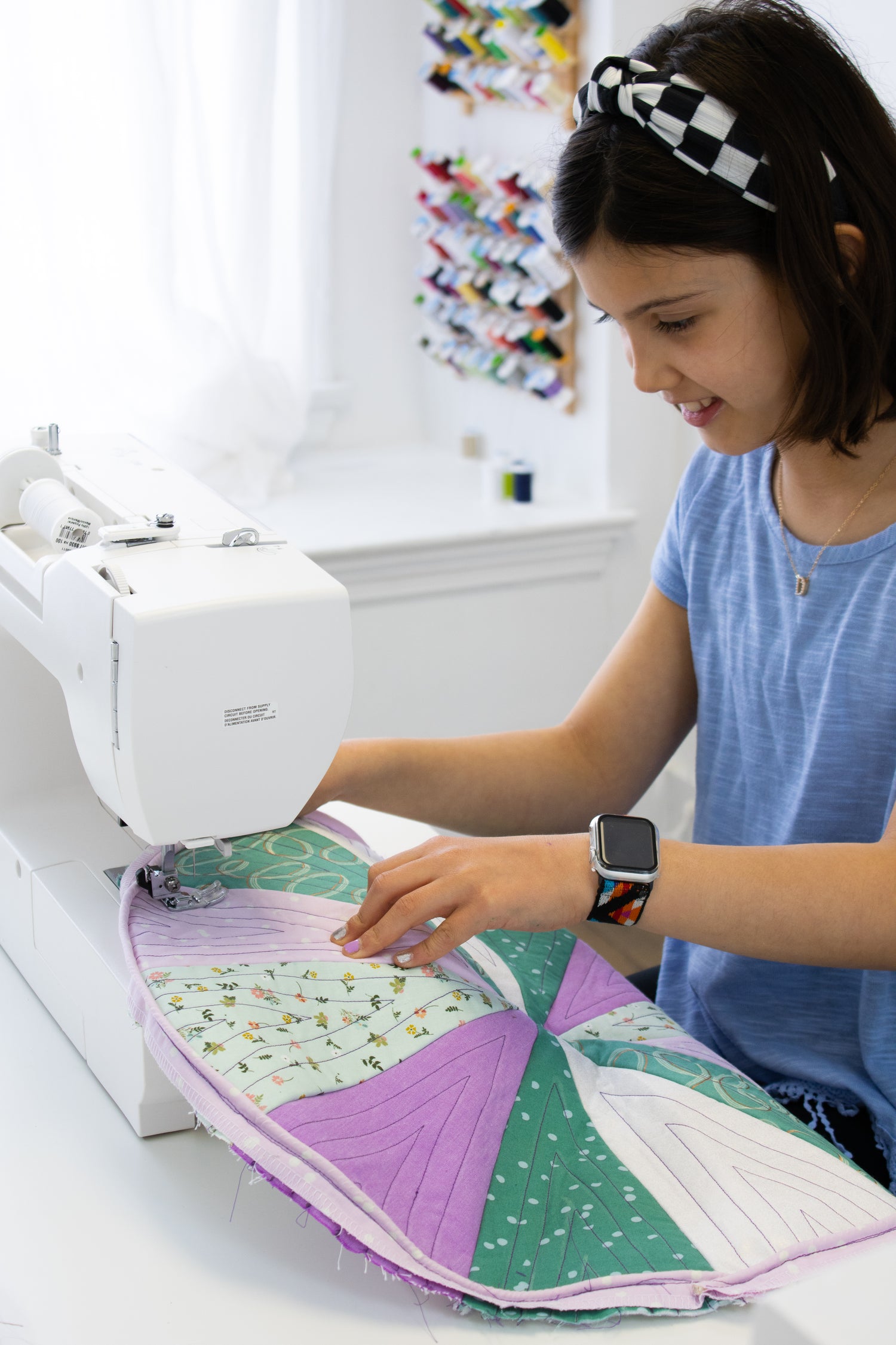 A girl wearing a headband and smartwatch uses a sewing machine to stitch a colorful quilted fabric piece in a bright room with spools of thread on the wall in the background.