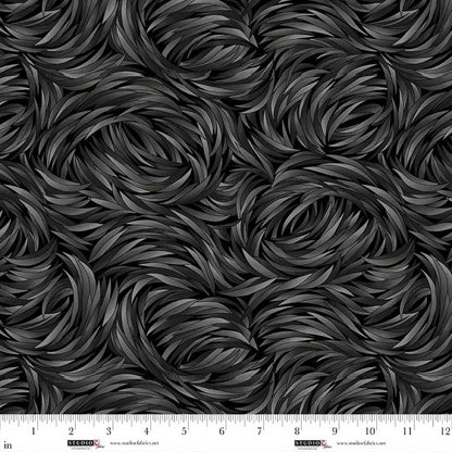 A close-up of QT Fabrics' Tempest Charcoal Swirl 108" Wide Quilt Backing Fabric shows a black and dark gray feather or leaf pattern, with a ruler along the bottom edge marking inches 1–13 to illustrate the fabric's scale.
