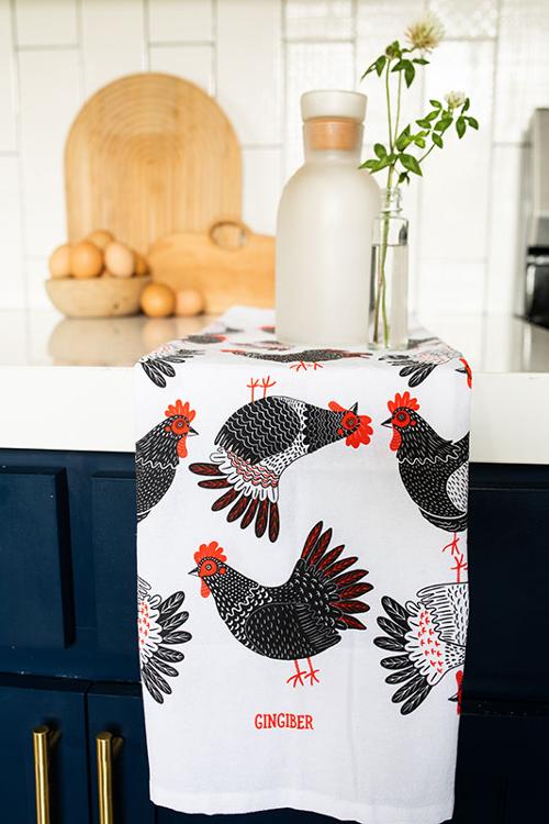A United Notions Gingiber Tea Towel Chicken with a black and red chicken print hangs over a dark blue cabinet. On the counter are a glass bottle, small vase with white flower, eggs, and a wooden cutting board.