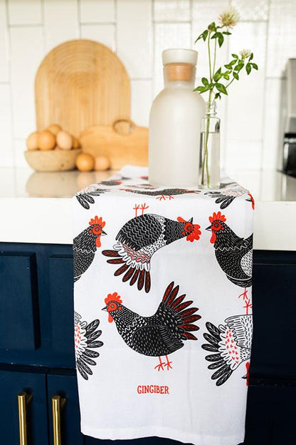 A United Notions Gingiber Tea Towel Chicken with a black and red chicken print hangs over a dark blue cabinet. On the counter are a glass bottle, small vase with white flower, eggs, and a wooden cutting board.